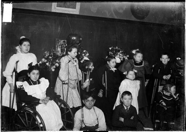 Children with various disabilities pose in front of a classroom blackboard. Some are in wheelchairs and some have crutches. The girls have long, puffy dresses on, the boys wear dress shirts and suspenders.